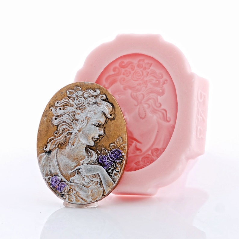 May include: An oval-shaped silicone mold in light pink featuring a recessed cameo design of a person with flowers. Beside it sits a finished cameo plaque in metallic gold and silver tones, depicting a person with purple floral accents.
