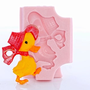 Könnte beinhalten: Eine gelbe Entenfigur mit roter Haube und Schal steht neben einer rosa Silikonform. Die Form zeigt eine Ente mit Haube und Schleife. Die Ente hat orangefarbene Füße und einen Schnabel. Die Figur ist etwa 5 cm hoch.