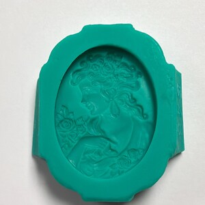May include: A teal silicone mold featuring an oval-shaped cameo design. The relief pattern depicts a profile of a person with wavy hair adorned with flowers, surrounded by a decorative border. The mold is flexible and has a smooth, matte finish.