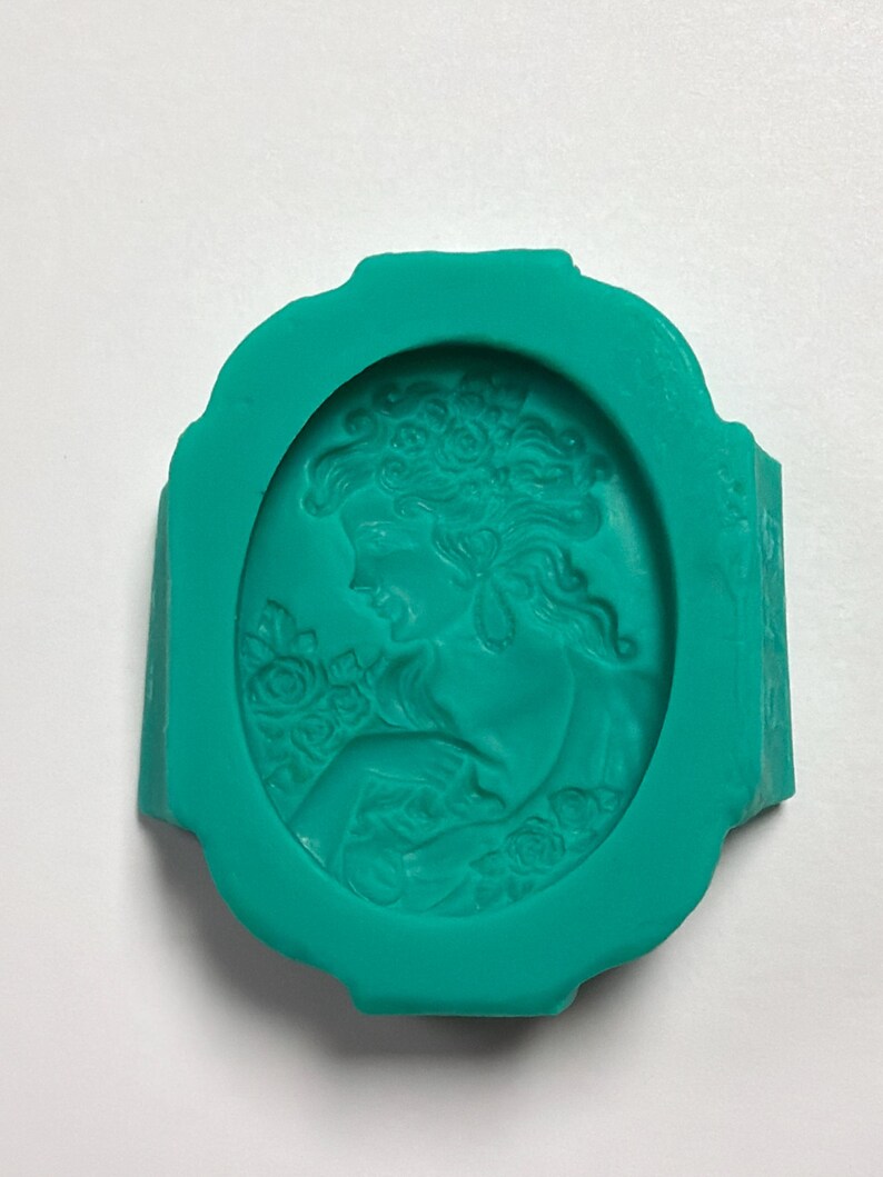 May include: A teal silicone mold featuring an oval-shaped cameo design. The relief pattern depicts a profile of a person with wavy hair adorned with flowers, surrounded by a decorative border. The mold is flexible and has a smooth, matte finish.