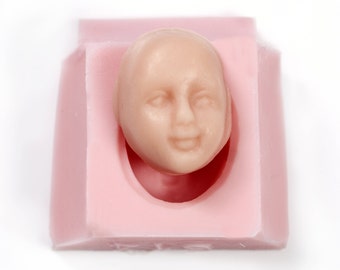 May include: A pink silicone mold with a detailed human face sculpted inside. The face is made of a light brown material and has a neutral expression.