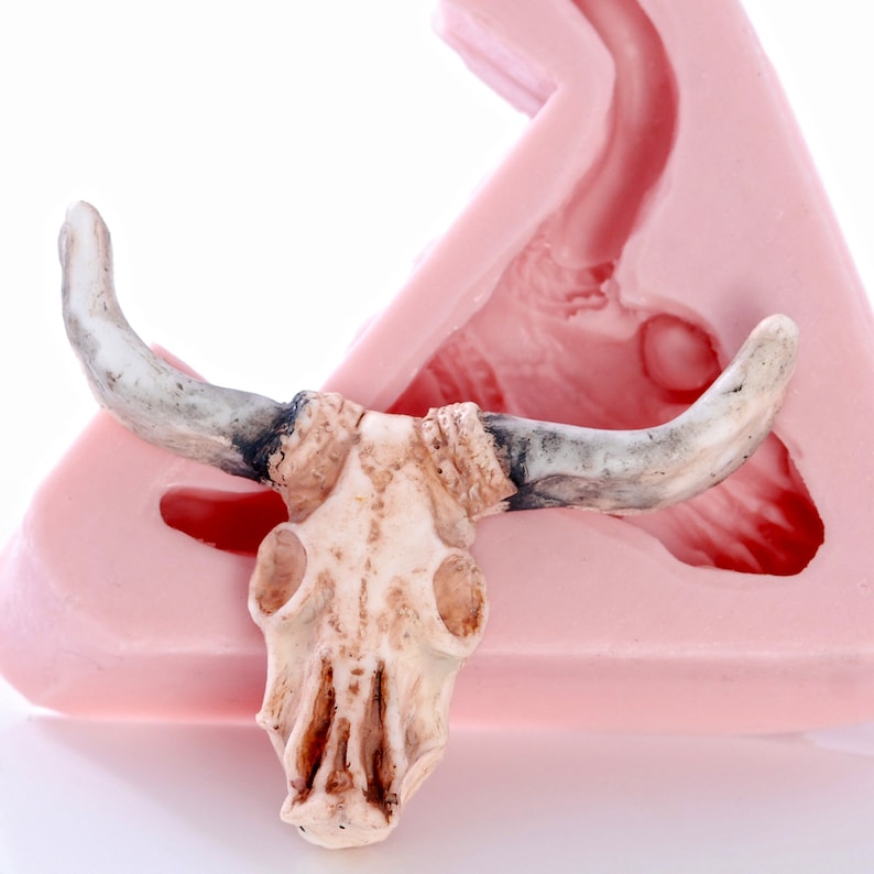May include: A white ceramic bull skull with horns, sitting on a pink silicone mold. The mold is shaped like a bull's head, with a hollow space for the skull to fit into.