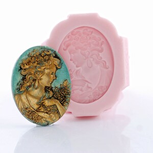 May include: An oval-shaped silicone mold in light pink featuring a relief of a person with flowing hair and flowers, accompanied by a finished oval cameo piece in teal and gold with the same detailed portrait design.