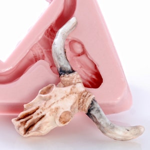 May include: A white ceramic bull skull with horns, resting on a pink silicone mold. The mold is shaped like a bull's head, with a detailed impression of the skull and horns.