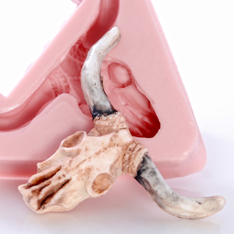 May include: A white ceramic bull skull with horns, resting on a pink silicone mold. The mold is shaped like a bull's head, with a detailed impression of the skull and horns.