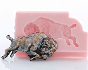 May include: A bronze-colored bison figurine with a detailed textured surface. The figurine is lying on its side with its head turned to the right. The figurine is next to a pink silicone mold of a bison.