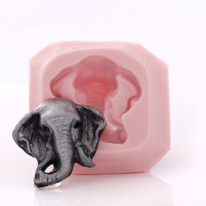 Puede incluir: Un molde de silicona rosa para hacer manualidades con forma de cabeza de elefante. Se muestra una cabeza de elefante gris junto al molde.
