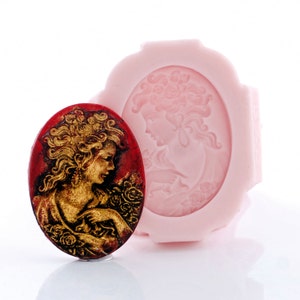 May include: An oval-shaped silicone mold in light pink featuring a relief of a person with flowing hair and roses, accompanied by a finished oval casting in deep red and metallic gold showing the same detailed portrait design.