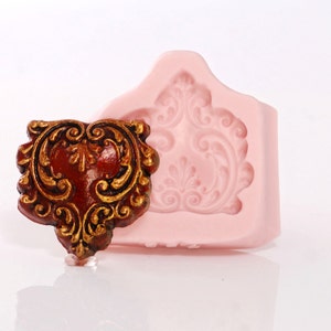 May include: A pink silicone mold with a detailed ornate design. The mold is used to create a decorative piece with a similar design. The piece is made of a red and gold material.