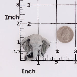 Puede incluir: Una figurita de cer&aacute;mica gris en forma de cabeza de elefante con una trompa y orejas detalladas. La figurita mide aproximadamente 2,5 cm de alto y se muestra sobre un fondo cuadriculado blanco con una regla.