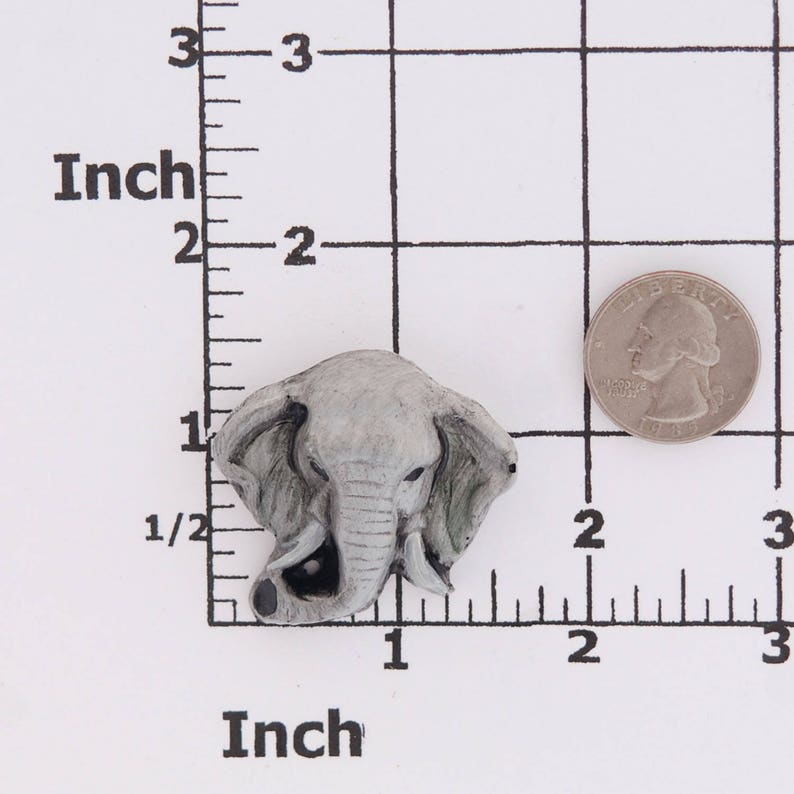Puede incluir: Una figurita de cer&aacute;mica gris en forma de cabeza de elefante con una trompa y orejas detalladas. La figurita mide aproximadamente 2,5 cm de alto y se muestra sobre un fondo cuadriculado blanco con una regla.