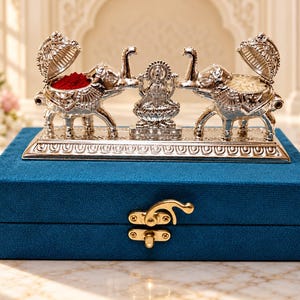 May include: A silver-toned metal figurine set featuring two elephants with hinged lids and a central deity figure, mounted on a rectangular base. The set rests atop a rectangular blue velvet box with a gold-colored metal latch closure.