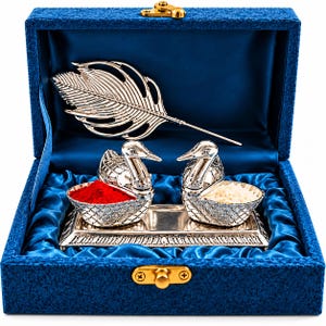 Puede incluir: Una caja forrada en terciopelo azul que contiene una bandeja de metal plateado, dos recipientes con forma de cisne y un objeto con forma de pluma. Un cisne contiene polvo rojo y el otro granos de arroz blanco. Cierre de metal dorado.