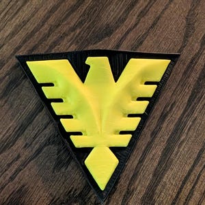May include: A triangular patch featuring a bright yellow eagle emblem centered on a black background. The eagle design is stylized with sharp, angular wings and a pointed tail, creating a high-contrast graphic element with a textured surface.