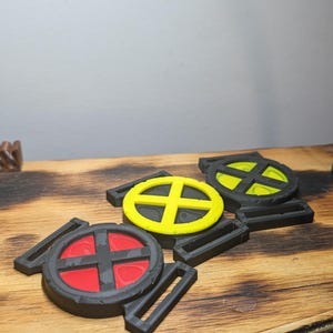 May include: Three circular black plastic buckles featuring a central cross design. One buckle has a red insert, one has a bright yellow insert, and the third has a lime green insert. Each buckle has rectangular slots on the sides for webbing.
