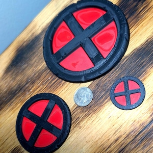 May include: Three circular emblems featuring a black frame with a raised black cross design over a bright red background. The set includes one large, one medium, and one small emblem, shown next to a silver US dime for scale reference.