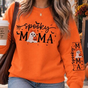 Peut inclure: Un sweat-shirt orange à manches longues avec le texte noir Spooky Mama, un motif de fantôme avec une citrouille et de petites illustrations de chauves-souris. Le même texte et motif sont répétés dans un cadre rectangulaire noir sur la manche.