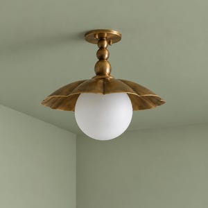 Può includere: Una plafoniera con base in metallo finitura ottone, uno stelo a sfere sovrapposte e un ampio paralume dai bordi smerlati. Un grande globo sferico in vetro bianco satinato è posizionato centralmente sotto il paralume in metallo.