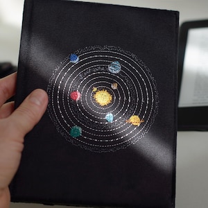 May include: Black book cover featuring an embroidered solar system design. The design includes a yellow sun at the center, surrounded by planets in various colors, and white dotted orbital paths. The cover is held by a hand.