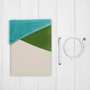 May include: A teal, green, and beige geometric pattern tablet case. A white pen and a white charging cable are next to the case. The case is made of a soft, velvety material.