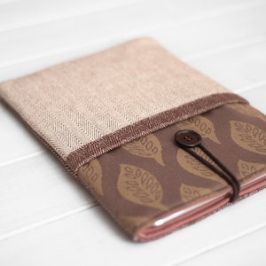 May include: A fabric book cover with a beige herringbone pattern top and a brown leaf-patterned bottom. A brown button and elastic closure secures the cover. The cover is made of soft fabric and is designed to protect books.