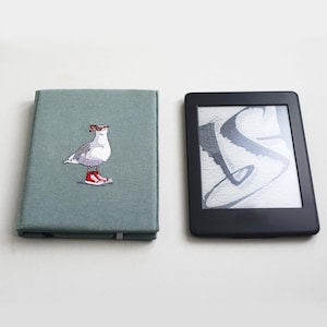 May include: A green fabric notebook cover with an embroidered image of a seagull wearing sunglasses and red shoes. The cover is next to a black e-reader.