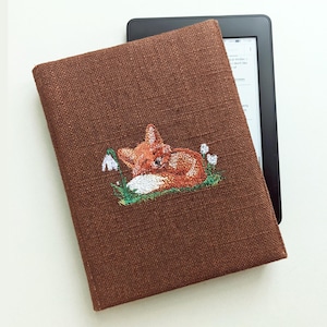 Op de afbeelding: Een bruine linnen e-reader hoes met een geborduurd ontwerp van een slapende vos. De vos is oranje en wit, rustend op groen gras met witte bloemen. De hoes beschermt een zwart e-reader apparaat.