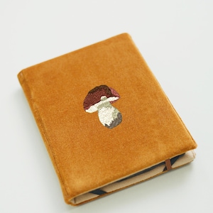 May include: A mustard-colored journal with an embroidered mushroom design. The mushroom has a brown cap, a white stem, and a textured appearance. The journal is rectangular and appears to be made of a soft, velvety material.