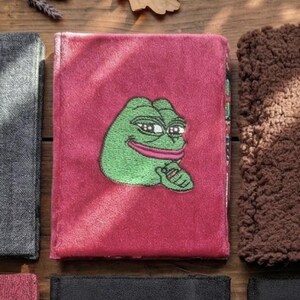 Könnte beinhalten: Ein roter, plüschiger Notizbuchumschlag mit einem gestickten grünen Froschcharakter mit einem lächelnden Ausdruck. Das Notizbuch ist von anderen Stoffartikeln in Grau-, Schwarz- und Brauntönen mit einem Holzuntergrund umgeben.