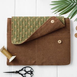 May include: A brown laptop sleeve with a green quilted flap secured by a button. The sleeve has a pocket and a button closure. A spool of thread and black scissors are next to the sleeve.