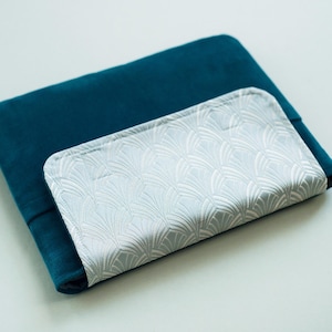 May include: A teal velvet cushion with a light blue, patterned rectangular object on top. The rectangular object has a repeating fan-like design. The cushion is folded over at the bottom.