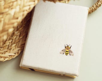 May include: A white notebook with a small embroidered bee design. The bee is yellow and black with brown wings.