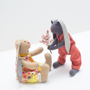 May include: Two plush bunny toys, one gray and one beige, are sitting on a white surface. The gray bunny is holding a bouquet of pink flowers and is looking at the beige bunny. The beige bunny is wearing a yellow and pink floral dress and is looking at the gray bunny.