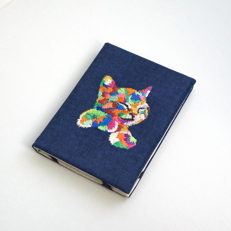 May include: A dark blue notebook with a colorful embroidered cat design. The cat is facing the right side of the notebook and has a happy expression.