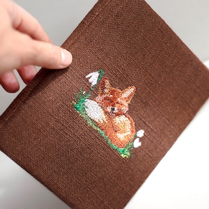 以下が含まれることがあります： 茶色のリネンの正方形で、眠っているキツネの刺繍デザインが施されています。キツネはオレンジと白で、白い花のある緑の草の上にいます。生地は手で持たれています。