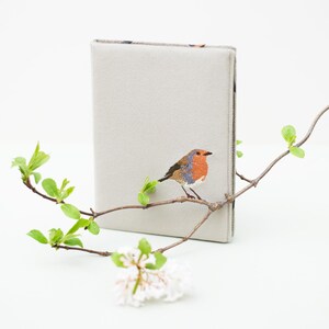 May include: A gray linen notebook cover with an embroidered robin perched on a branch. The cover has a subtle texture and a simple design.