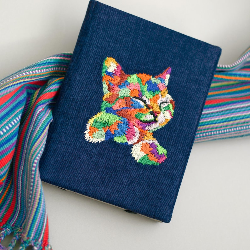 May include: A dark blue denim notebook with a colorful embroidered cat design. The cat is facing right and has a playful expression.