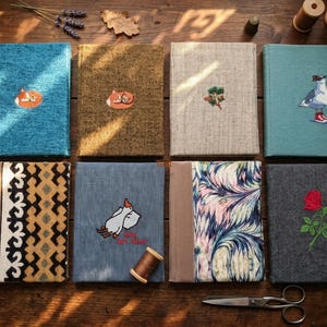 May include: Eight fabric-covered journals with embroidered designs, including a fox, a seagull, and a rose. The journals are in various colours and patterns, arranged on a wooden surface with sewing supplies. The journals are of various sizes.