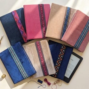 Könnte beinhalten: Eine Sammlung von sieben rechteckigen Stoffhüllen in verschiedenen Farben wie Marineblau, Pink, Beige und Mauve, die jeweils mit einer vertikalen, dekorativen Webborte versehen sind. Ein Nahttrenner, ein Fingerhut und Fäden liegen daneben.
