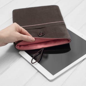 May include: A brown felt tablet sleeve with a pink lining and a button closure. The sleeve is open and a white tablet is inside.