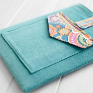 May include: A teal-colored, rectangular fabric item with a smaller, lighter teal pocket. A patterned fabric strip with orange, blue, and pink floral designs is attached. The item appears to be a case or cover.