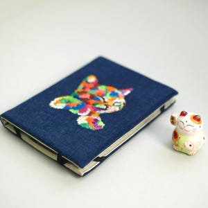 May include: A blue denim notebook with a colorful embroidered cat design. The notebook is closed and lying on a white surface. A small white ceramic cat figurine is sitting to the right of the notebook.
