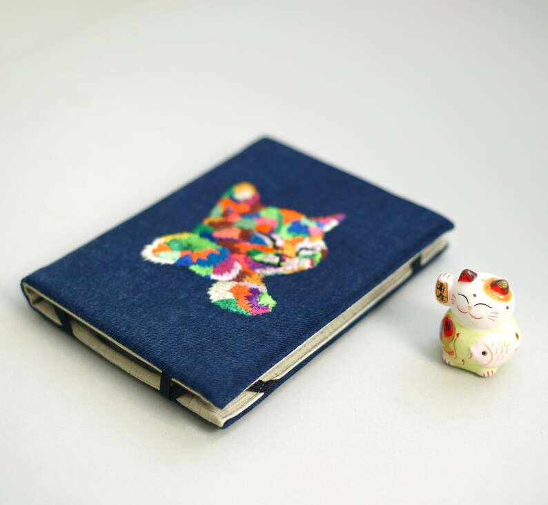 May include: A blue denim notebook with a colorful embroidered cat design. The notebook is closed and lying on a white surface. A small white ceramic cat figurine is sitting to the right of the notebook.