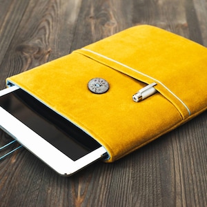 Peut inclure: Une housse de tablette jaune moutarde en velours doux. La housse a une bordure bleu clair et une fermeture à bouton décorative. Un stylo argenté est glissé dans une poche latérale. Une tablette blanche est partiellement visible.