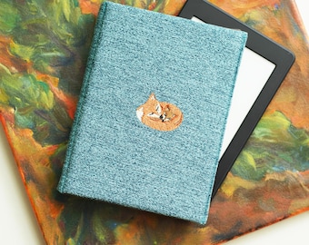 May include: A teal blue notebook with a small embroidered fox design. The notebook is lying on a colorful abstract background.