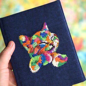 May include: A dark blue denim passport holder with a colorful embroidered cat design. The cat is depicted with its eyes closed and a playful expression.