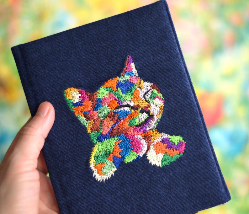 May include: A dark blue denim passport holder with a colorful embroidered cat design. The cat is depicted with its eyes closed and a playful expression.