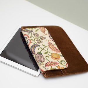 May include: A brown suede tablet case with a floral patterned flap. The case is open and a tablet is inside.