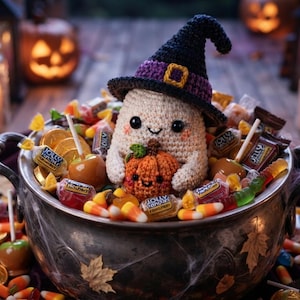 May include: A crocheted ghost wearing a black and purple witch hat, holding a small crocheted orange pumpkin. The ghost sits inside a metallic cauldron filled with assorted sweets, including candy corn, wrapped chocolates, and caramel apples.