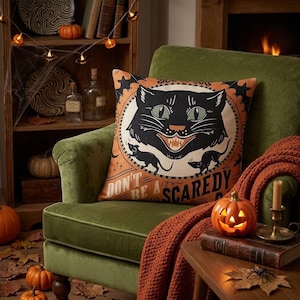 May include: A square throw pillow featuring a black cat illustration with green eyes and sharp teeth, surrounded by small black mice and bats on an orange background. The text reads, "DON'T BE A SCAREDY CAT."